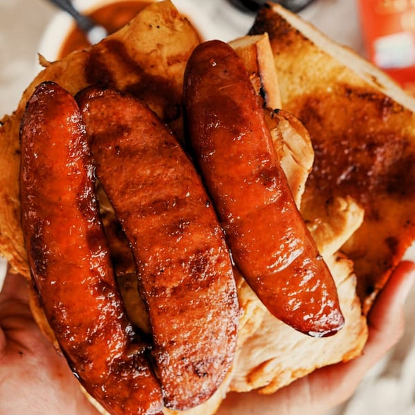 Close-up of two hands holding a Best BBQ sausage sandwich with grilled bread and glazed sausages.