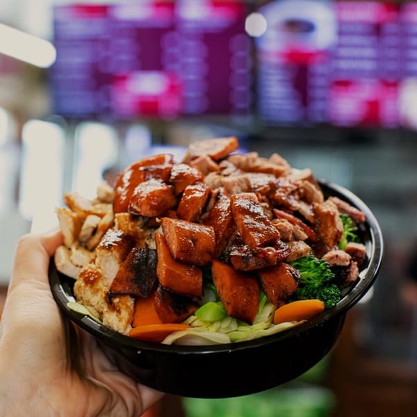A hand holds a Best BBQ bowl filled with chicken, beef, sweet potatoes, and vegetables.