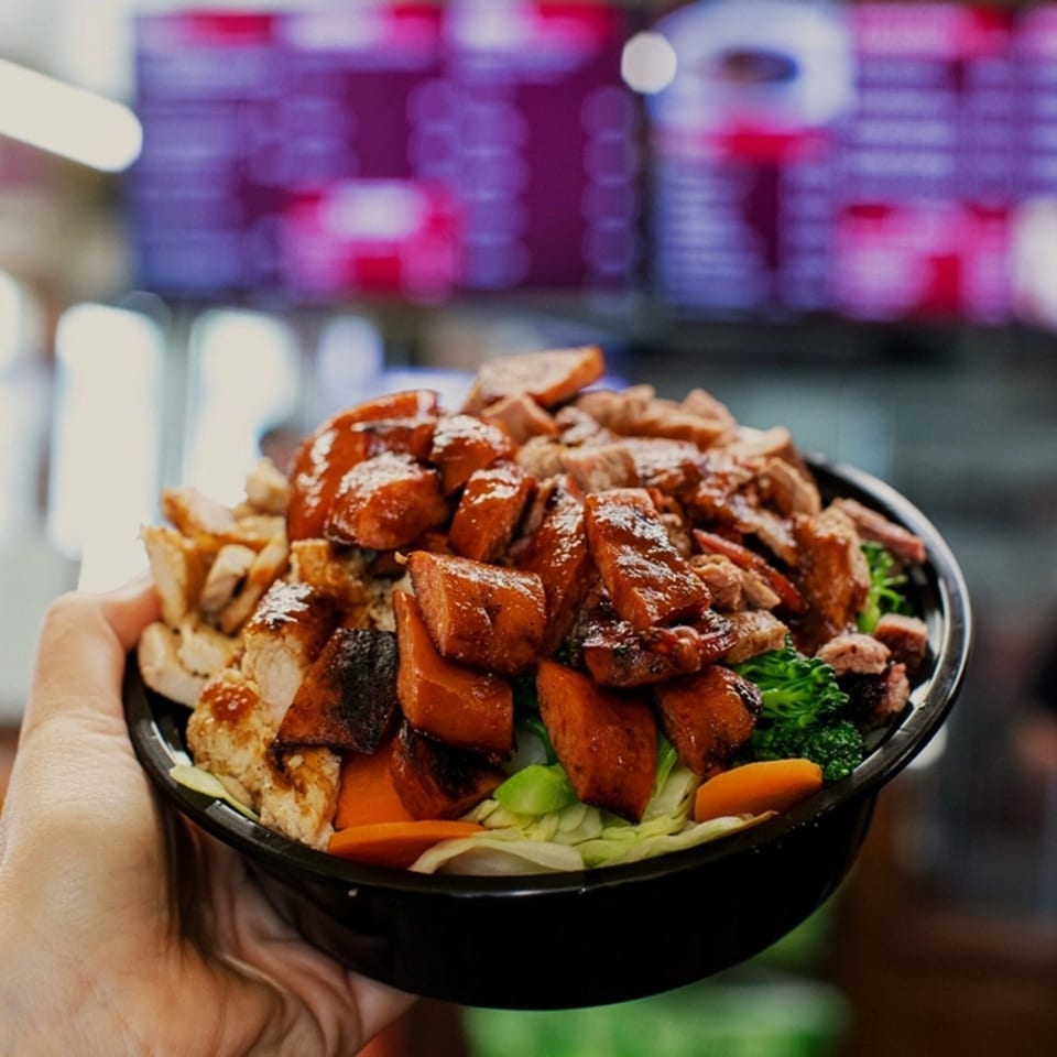 A hand holds a Best BBQ bowl filled with chicken, beef, sweet potatoes, and vegetables.