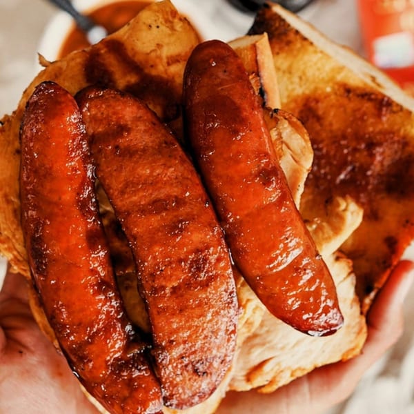 Three grilled sausages with barbecue sauce on toasted bread, held by a hand.