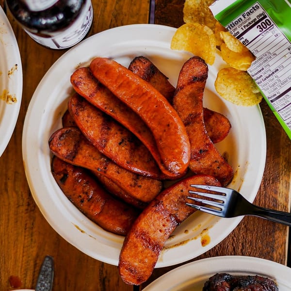 A plate of Best BBQ's grilled sausages with a fork, chips, and a drink on a wooden table.